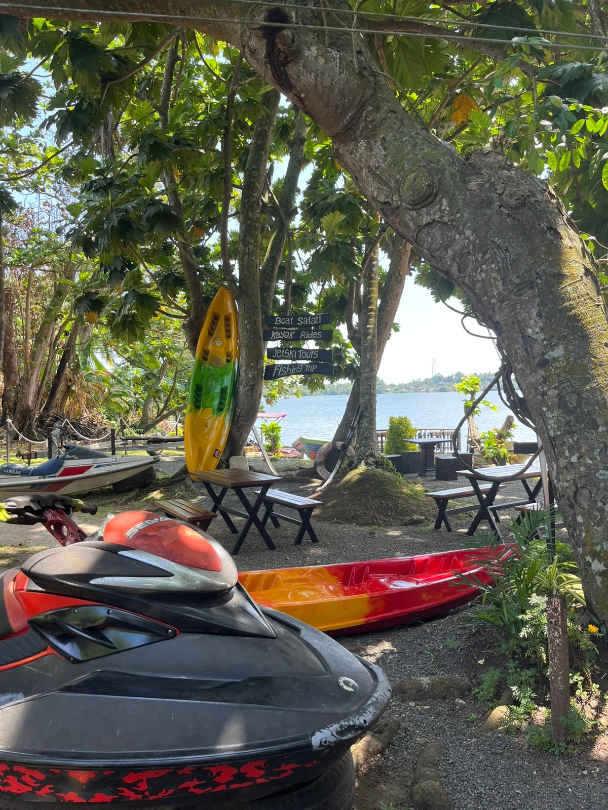 Shaded wooden picnic tables on the gravel riverbank beside the jetty, with boats moored on the Bentota River